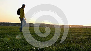 Young caucasian man in yellow jacket with backpack walking on meadow, field at sunset in czech spring landscape