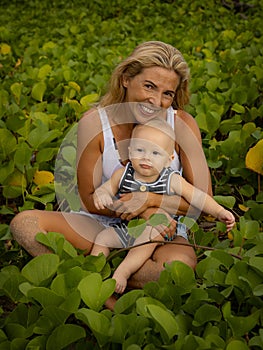 Young Caucasian mother with baby infant boy spending time in nature. Outdoor activities. Mom and baby boy sitting on the ground.