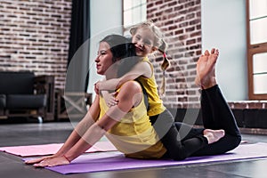 Young Caucasian mother doing yoga cobra pose on floor while her smiling daughter sitting on moms back hugging her in