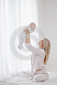 A young Caucasian mother in home clothes holds a newborn baby in a white jumpsuit. Smiles and hugs the child.