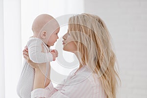 A young Caucasian mother in home clothes holds a newborn baby in a white jumpsuit. Smiles and hugs the child.