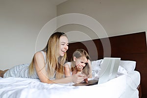Young caucasian mother lying in bed with little daughter and using laptop.
