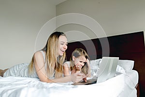 Young caucasian mother lying in bed with little daughter and using laptop.