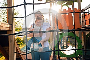 A young caucasian mother plays with a baby on the playground. Sunset in the background. Summertime