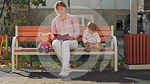 Young caucasian mother is reading a book sitting on a bench. A child is sitting nearby and watching cartoons on a