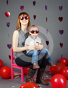 Young Caucasian mother sitting on small pink chair with baby boy toddler on her laps knees in studio wearing funny glasses
