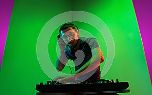 Young caucasian musician in headphones performing on bicolored green-purple background in neon light