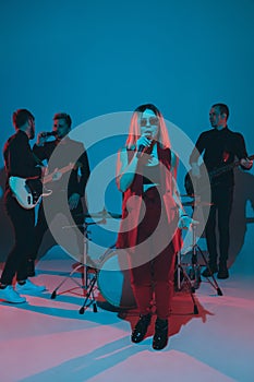 Young caucasian musicians, band performing in neon light on blue studio background, singer in front