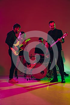 Young caucasian musicians, band performing in neon light on red studio background