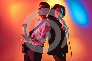 Young caucasian musicians, female singer and guitarist performing on gradient background in neon light. Concept of music
