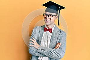 Young caucasian nerd man wearing glasses and graduation cap happy face smiling with crossed arms looking at the camera