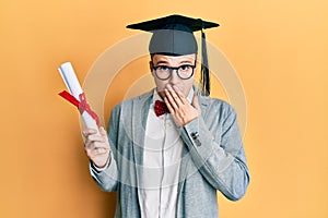 Young caucasian nerd man wearing glasses and graduation cap and holding degree covering mouth with hand, shocked and afraid for