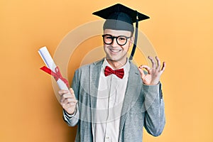 Young caucasian nerd man wearing glasses and graduation cap and holding degree doing ok sign with fingers, smiling friendly