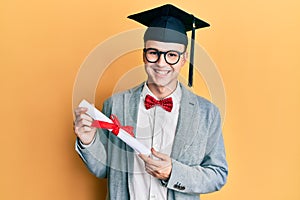 Young caucasian nerd man wearing glasses and graduation cap and holding degree looking positive and happy standing and smiling