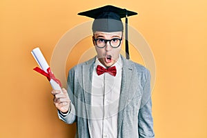 Young caucasian nerd man wearing glasses and graduation cap and holding degree scared and amazed with open mouth for surprise,