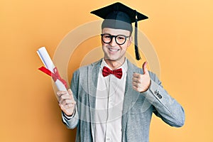 Young caucasian nerd man wearing glasses and graduation cap and holding degree smiling happy and positive, thumb up doing