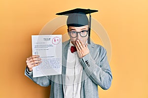 Young caucasian nerd man wearing glasses and graduation cap holding passed exam covering mouth with hand, shocked and afraid for