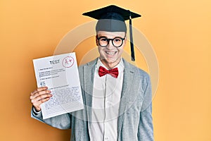 Young caucasian nerd man wearing glasses and graduation cap holding passed exam looking positive and happy standing and smiling