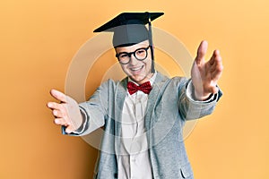 Young caucasian nerd man wearing glasses and graduation cap looking at the camera smiling with open arms for hug