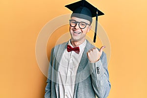 Young caucasian nerd man wearing glasses and graduation cap pointing thumb up to the side smiling happy with open mouth