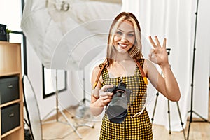 Young caucasian photographer girl holding professional camera at photography studio doing ok sign with fingers, smiling friendly