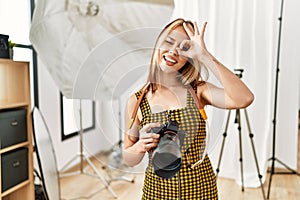 Young caucasian photographer girl holding professional camera at photography studio smiling happy doing ok sign with hand on eye