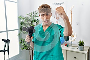 Young caucasian physio man holding muscle percusion gun at the clinic annoyed and frustrated shouting with anger, yelling crazy