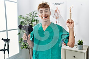 Young caucasian physio man holding muscle percusion gun at the clinic celebrating victory with happy smile and winner expression