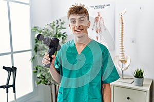 Young caucasian physio man holding muscle percusion gun at the clinic looking positive and happy standing and smiling with a