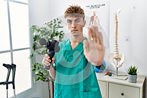 Young caucasian physio man holding muscle percusion gun at the clinic with open hand doing stop sign with serious and confident