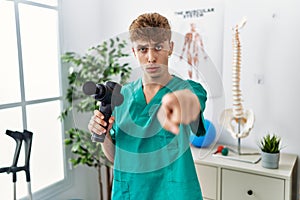 Young caucasian physio man holding muscle percusion gun at the clinic pointing with finger to the camera and to you, confident