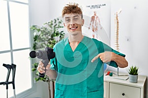 Young caucasian physio man holding muscle percusion gun at the clinic pointing finger to one self smiling happy and proud