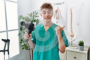 Young caucasian physio man holding muscle percusion gun at the clinic pointing thumb up to the side smiling happy with open mouth