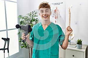 Young caucasian physio man holding muscle percusion gun at the clinic screaming proud, celebrating victory and success very
