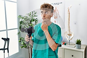 Young caucasian physio man holding muscle percusion gun at the clinic serious face thinking about question with hand on chin,