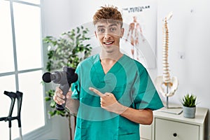 Young caucasian physio man holding muscle percusion gun at the clinic smiling happy pointing with hand and finger