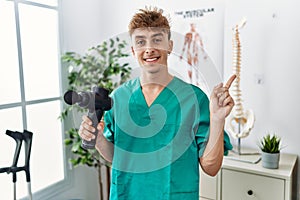 Young caucasian physio man holding muscle percusion gun at the clinic smiling happy pointing with hand and finger to the side
