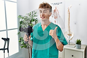 Young caucasian physio man holding muscle percusion gun at the clinic smiling happy and positive, thumb up doing excellent and