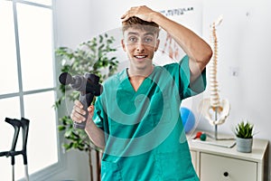 Young caucasian physio man holding muscle percusion gun at the clinic stressed and frustrated with hand on head, surprised and