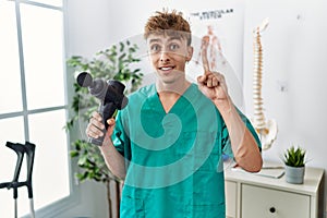 Young caucasian physio man holding muscle percusion gun at the clinic surprised with an idea or question pointing finger with