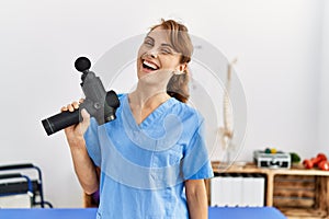 Young caucasian physio therapist girl smiling happy holding gun percusion at the clinic