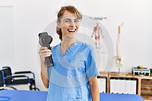 Young caucasian physio therapist girl smiling happy holding gun percusion at the clinic