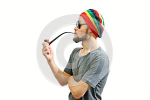 Young caucasian rasta man in jamaica hat, sunglasses and grey t-shirt on white background with smoke pipe in hand