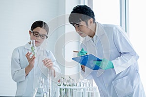 Young Caucasian scientist child girl hold flask with plant tissue and explain to her Asian teacher with record some data during do