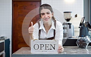 Young caucasian shopkeeper with a smile, lift a finger thumb up while holding an OPEN sign in front of a coffee shop counter