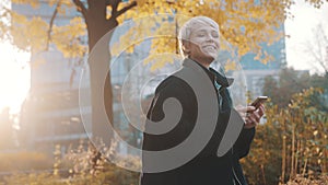 Young caucasian short haired blond woman using phone in the city park in front of office building in autumn
