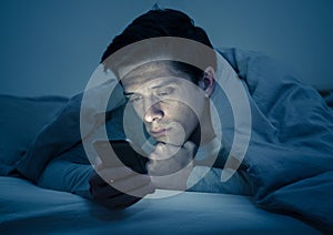 Young caucasian sleepless man bored in bed surfing on the Internet addicted to mobile phone