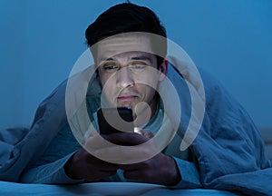 Young caucasian sleepless man bored in bed surfing on the Internet addicted to mobile phone