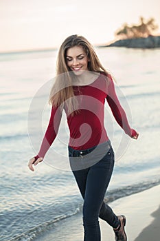 Young Caucasian slim woman with messy long hair wearing black jeans and red shirt walking on windy day outdoor on beach