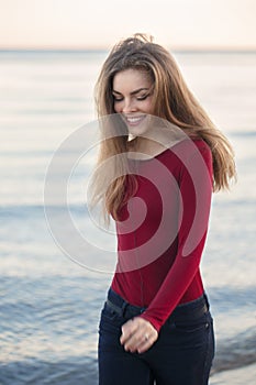 Young Caucasian slim woman with messy long hair wearing black jeans and red shirt walking on windy day outdoor on beach